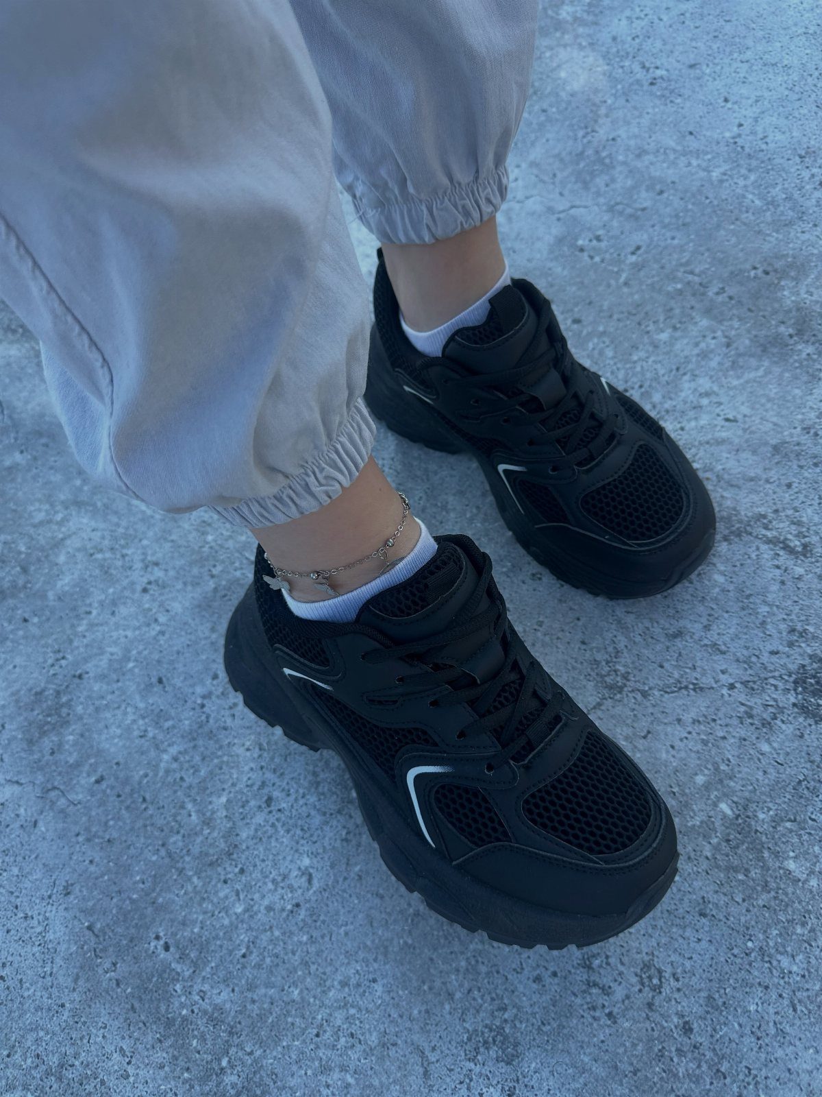 Black mesh athletic sneakers worn with light grey joggers on concrete ground.