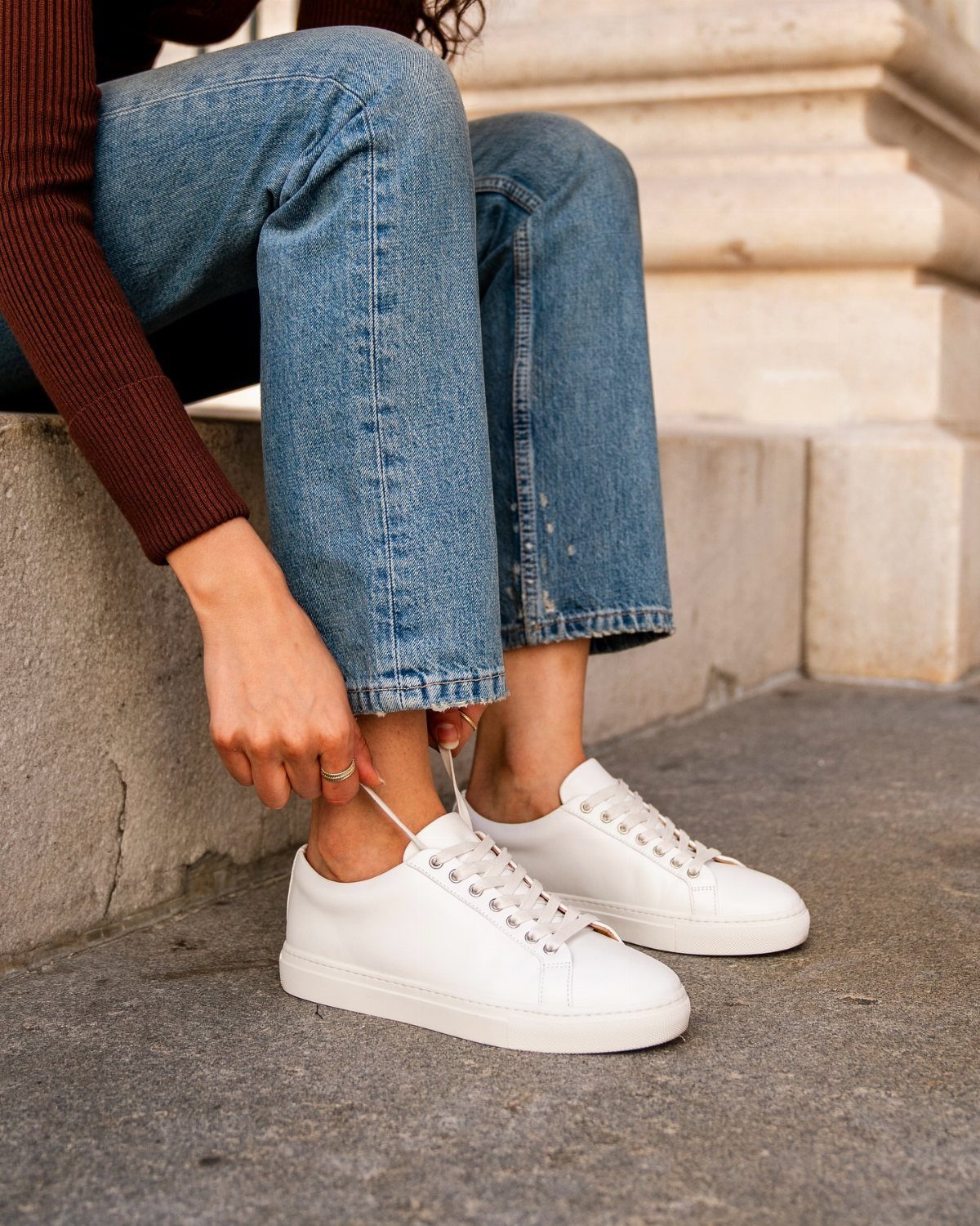 White leather low-top sneakers worn with blue jeans on stone steps.