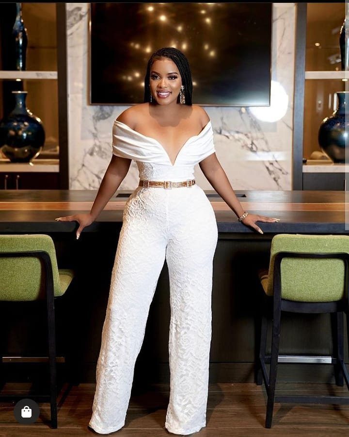 Woman in a white off-the-shoulder lace jumpsuit with a gold belt standing indoors.