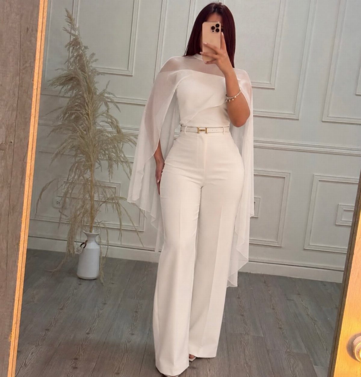 Woman in a white wide-leg jumpsuit with a sheer cape and thin belt.