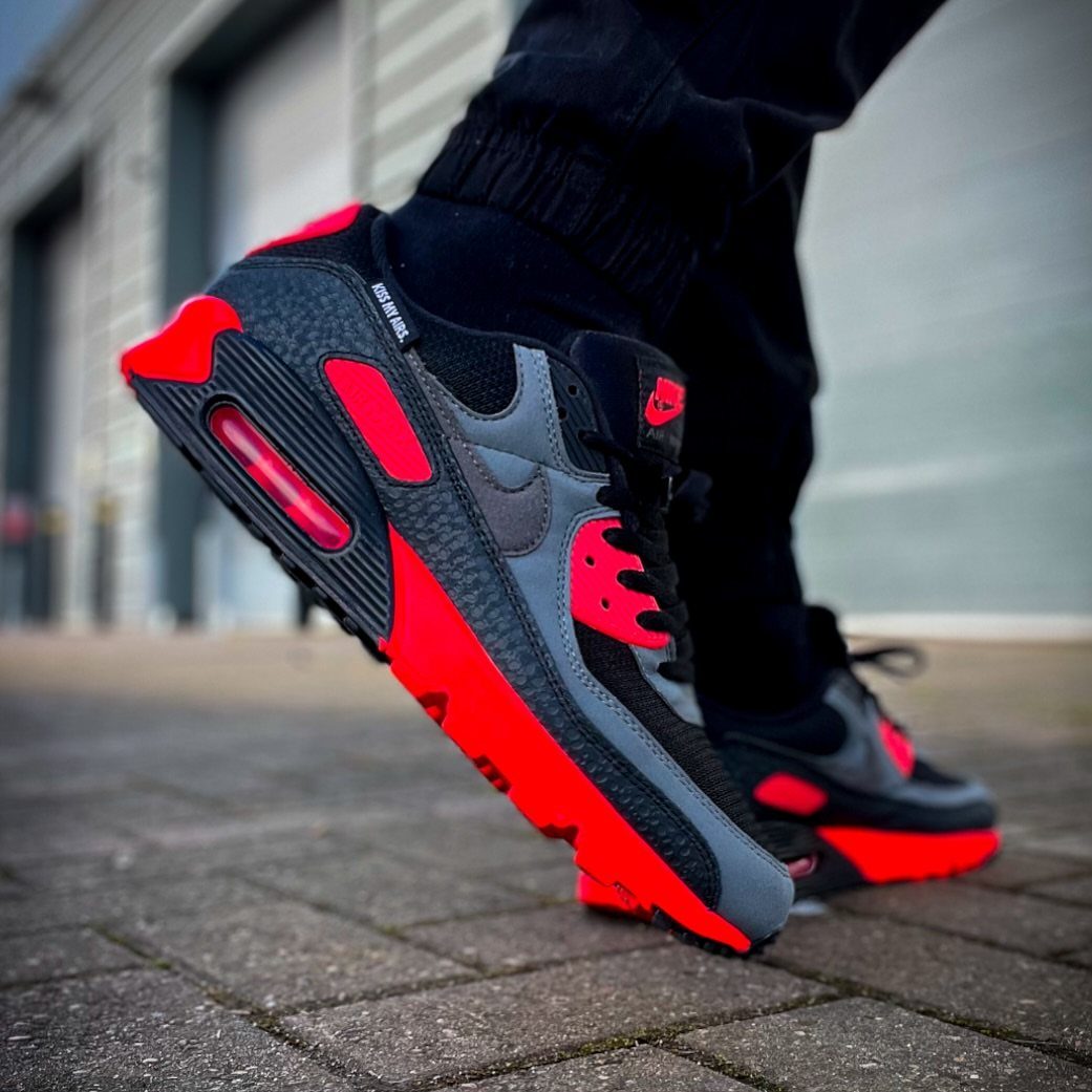 Red and black Nike Air Max 90 sneakers worn on a brick pavement.