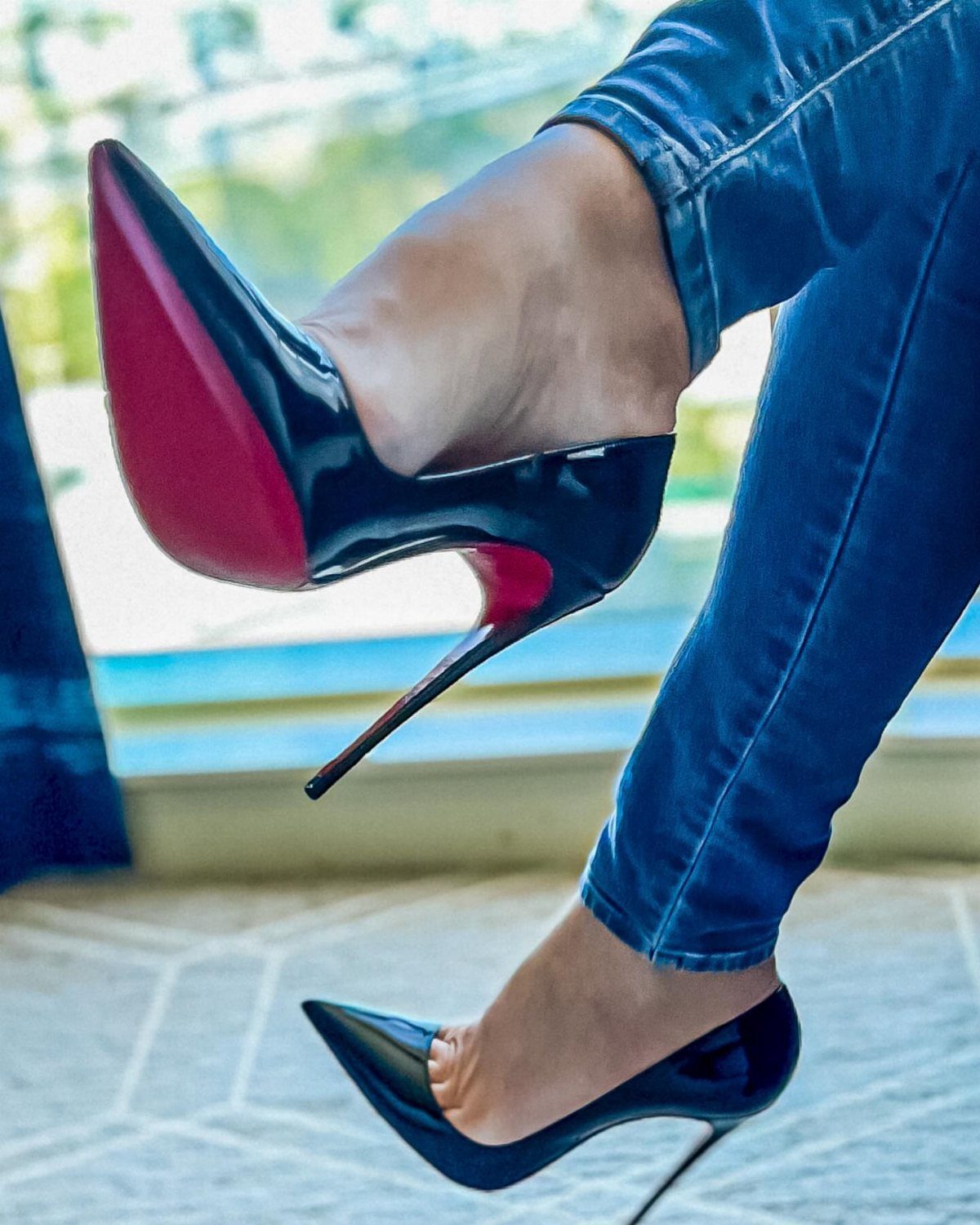 Black patent leather high heels with red soles worn with blue jeans.
