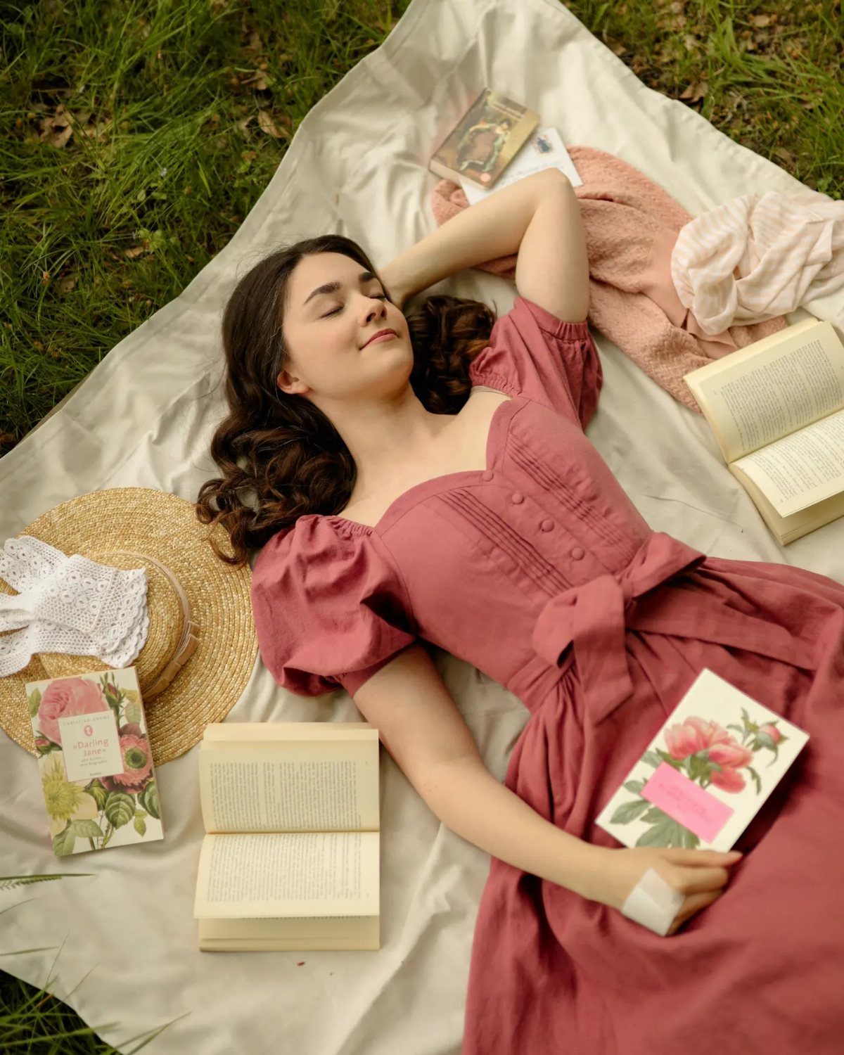 Young woman in a rose dress lying on a blanket with books and hat.