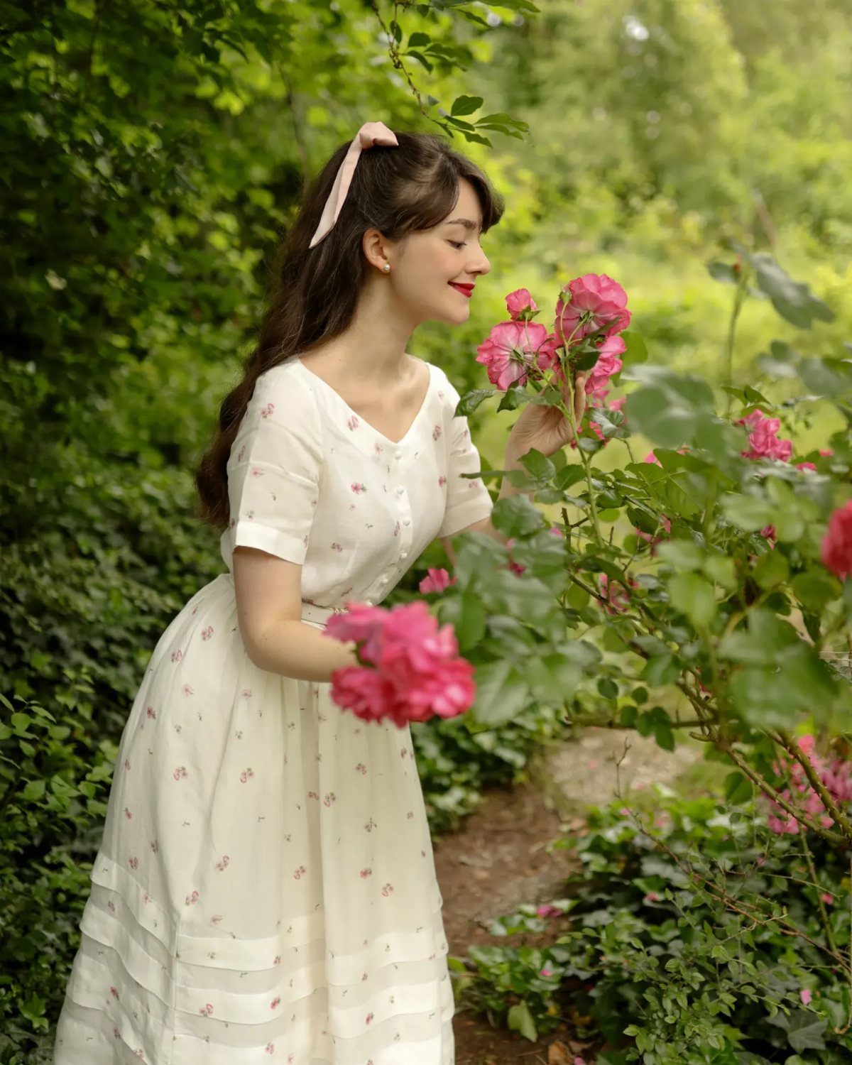 Woman in a white dress with small floral prints standing among pink roses.