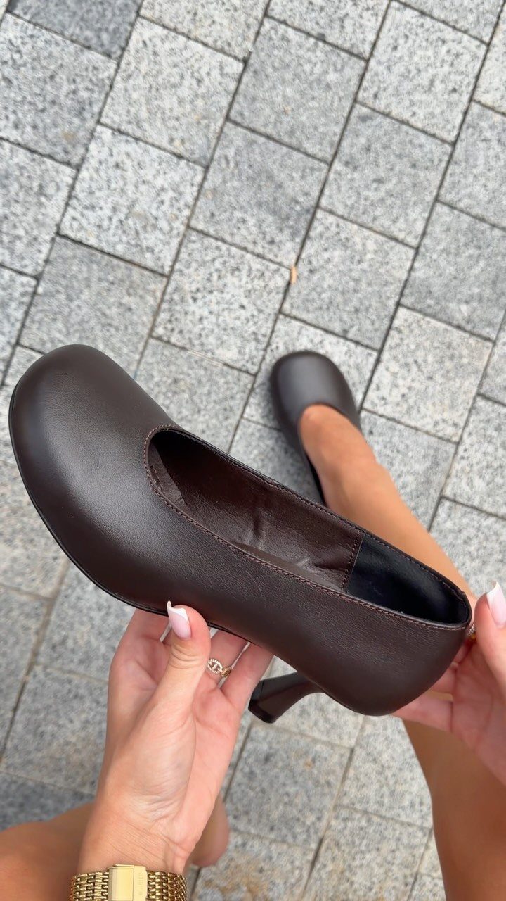 Brown round toe kitten heels held by a person over cobblestone pavement
