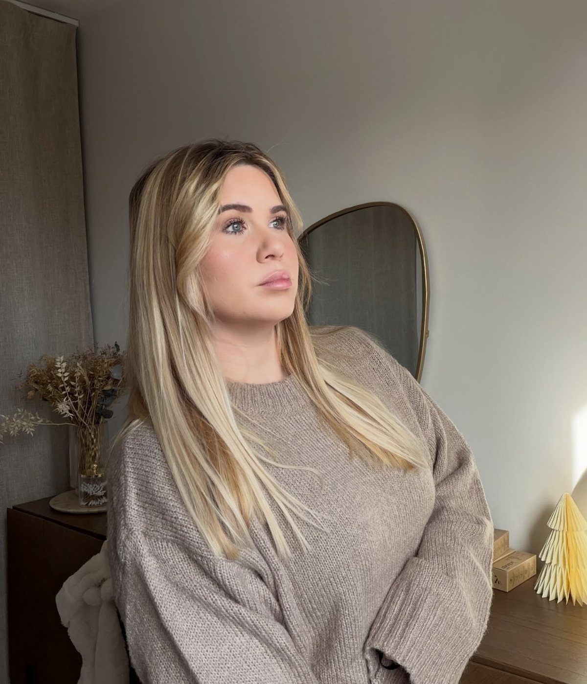 9 Soft Femme Style Ideas with Ruffles, Knits and Pearls 8 Woman with blonde hair wearing a cozy beige knit sweater in a neutral room.