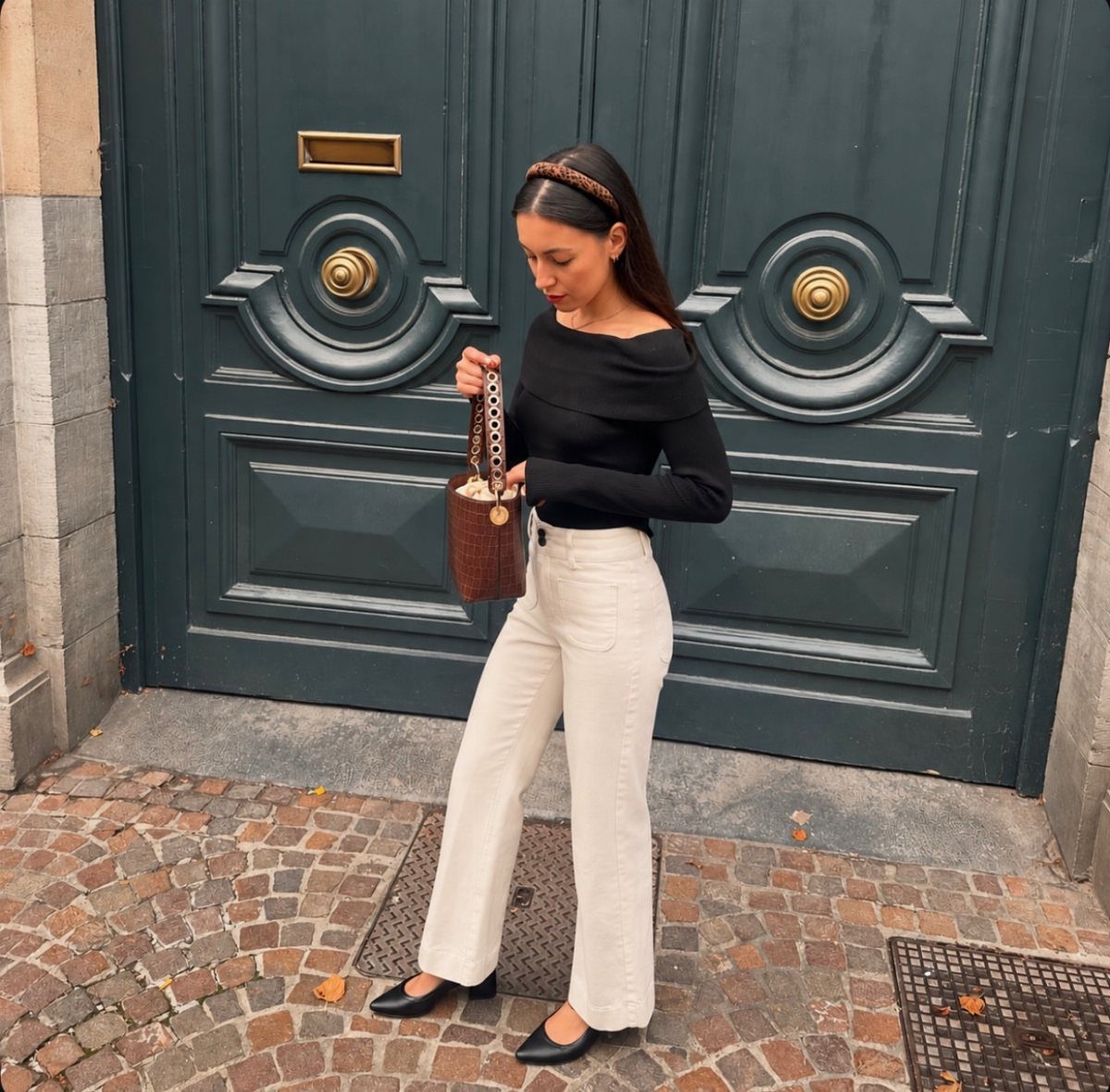9 Soft Femme Style Ideas with Ruffles, Knits and Pearls 7 Woman in black off-the-shoulder top and white trousers posing by a green door.