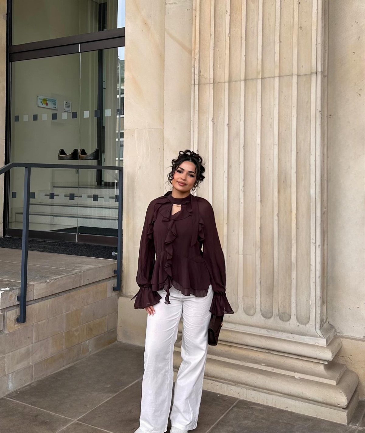 9 Soft Femme Style Ideas with Ruffles, Knits and Pearls 6 Woman in a burgundy ruffled blouse and white wide-leg trousers by a stone column.