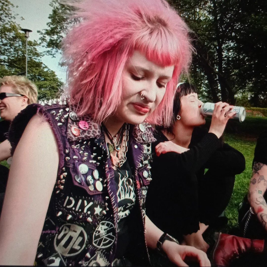 Person with pink spiky hair wearing a studded purple punk vest and patches.