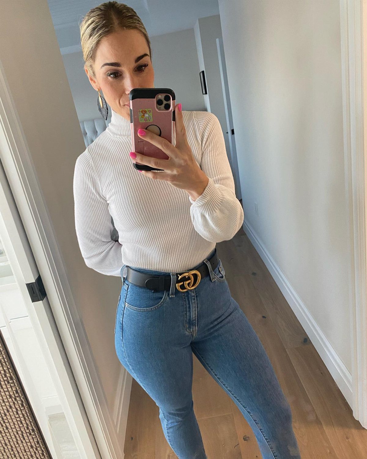 Woman in white turtleneck and blue high-waisted jeans taking a mirror selfie.