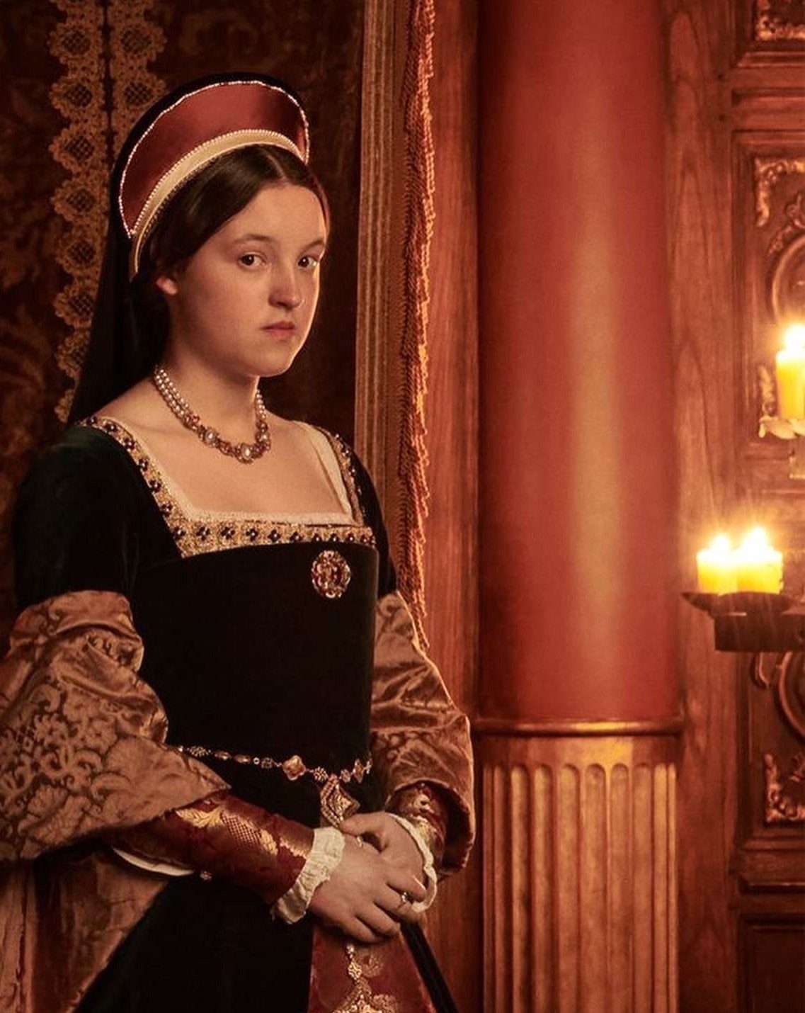 Woman in a green velvet Tudor gown and red French hood standing indoors.