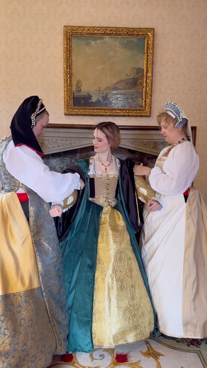 Three women wearing elaborate Tudor period dresses in a classically decorated room.