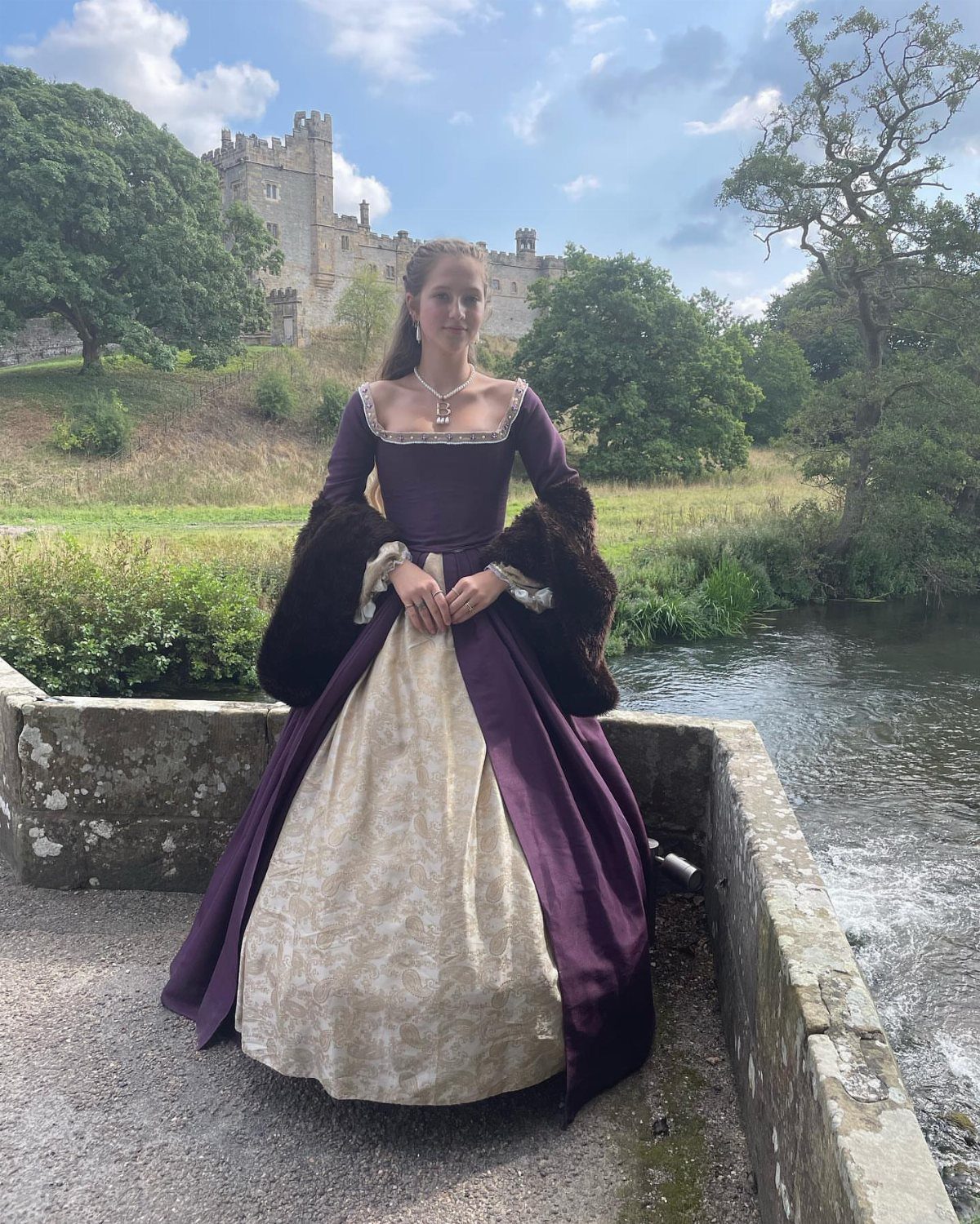 Woman in a purple and cream Tudor period dress standing before a castle.