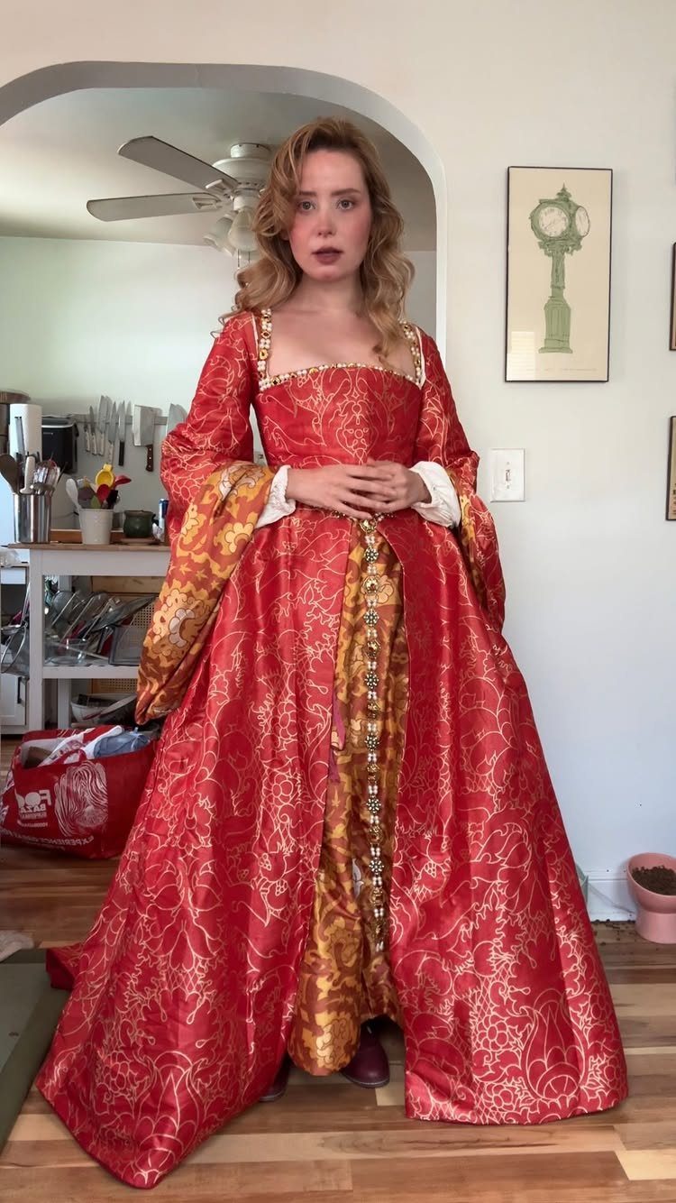 Woman wearing a red and gold brocade Tudor period dress in a room.