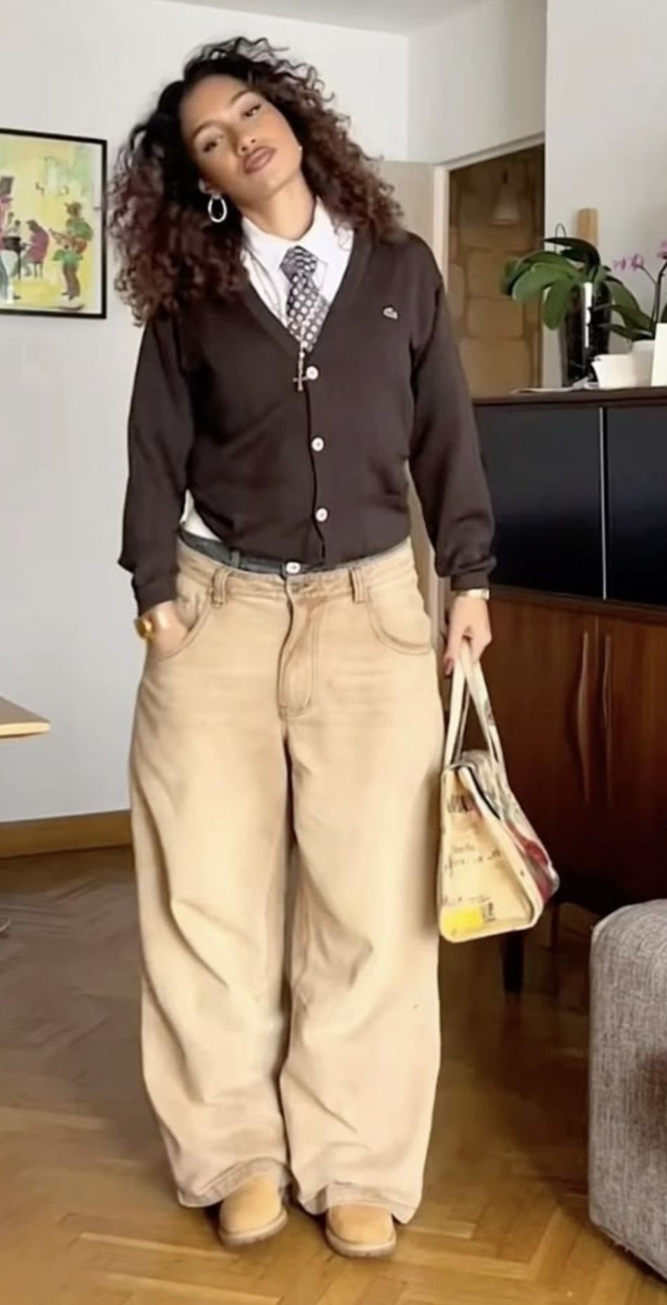 Woman wearing a brown cardigan, tie, and wide-leg tan trousers in a room.