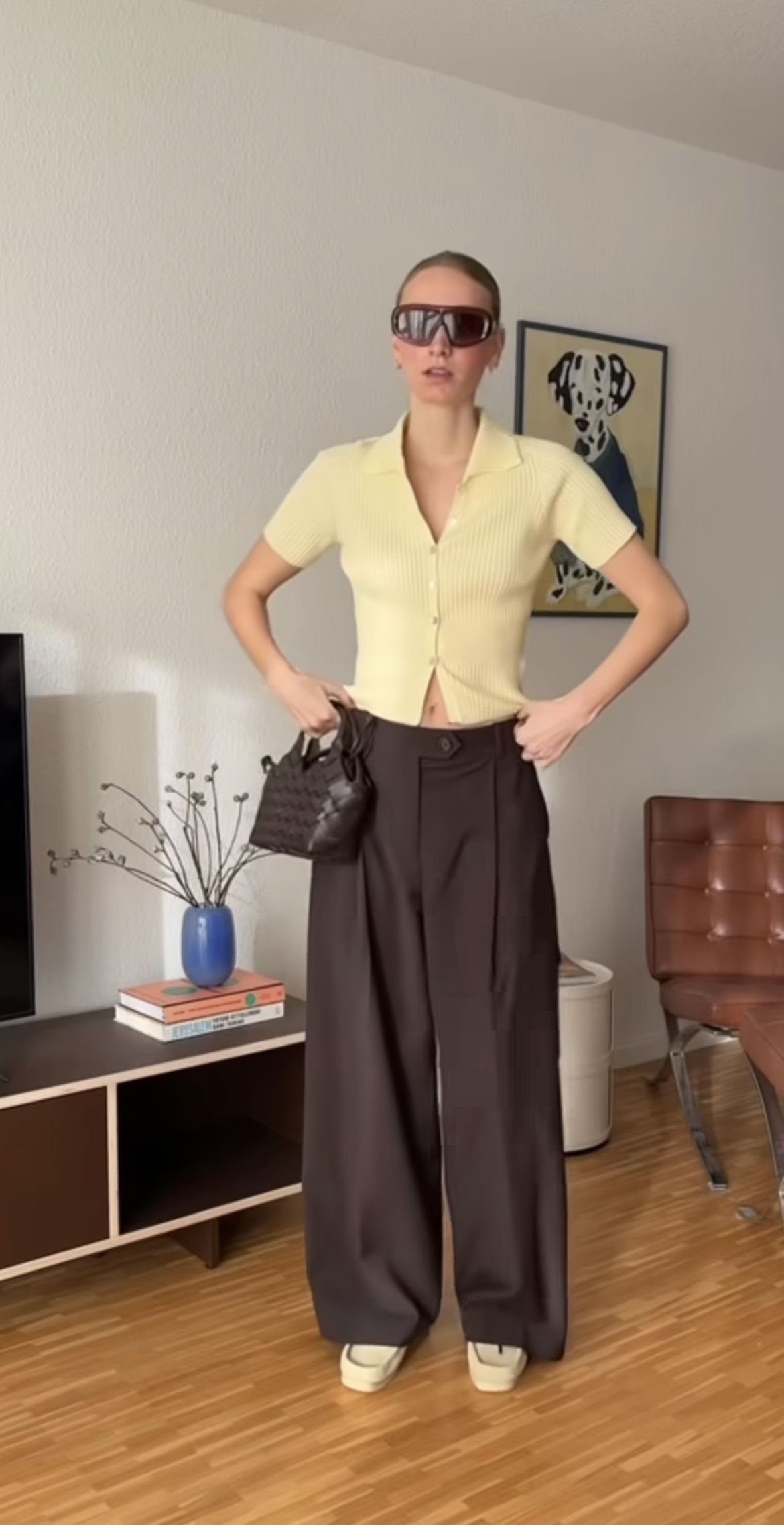 Woman wearing a pale yellow polo shirt and dark brown pleated wide-leg trousers.