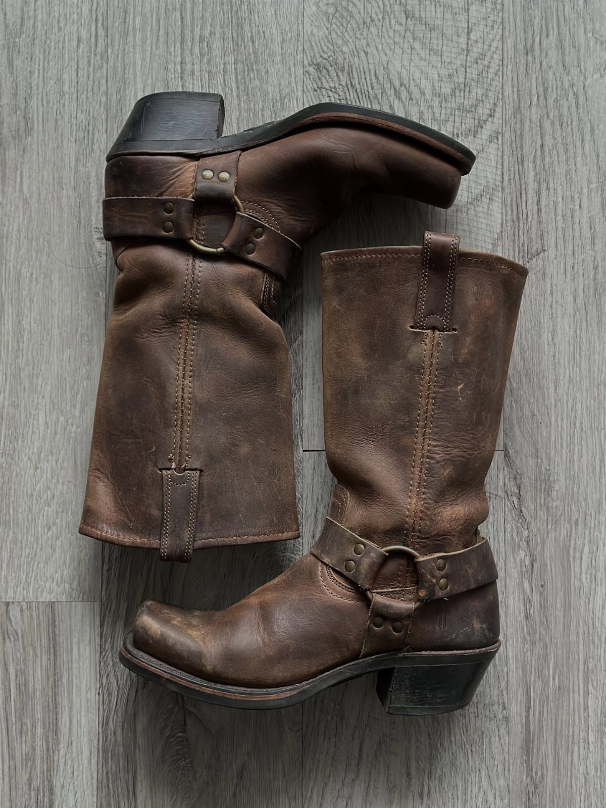Brown leather cowboy boots with buckles resting on a grey wooden floor.
