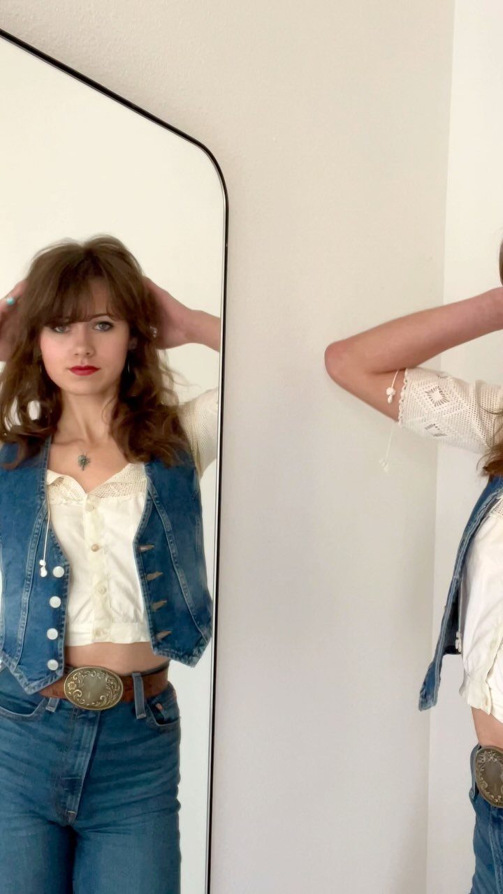 Woman wearing a cropped denim vest, white shirt, and belted blue jeans.