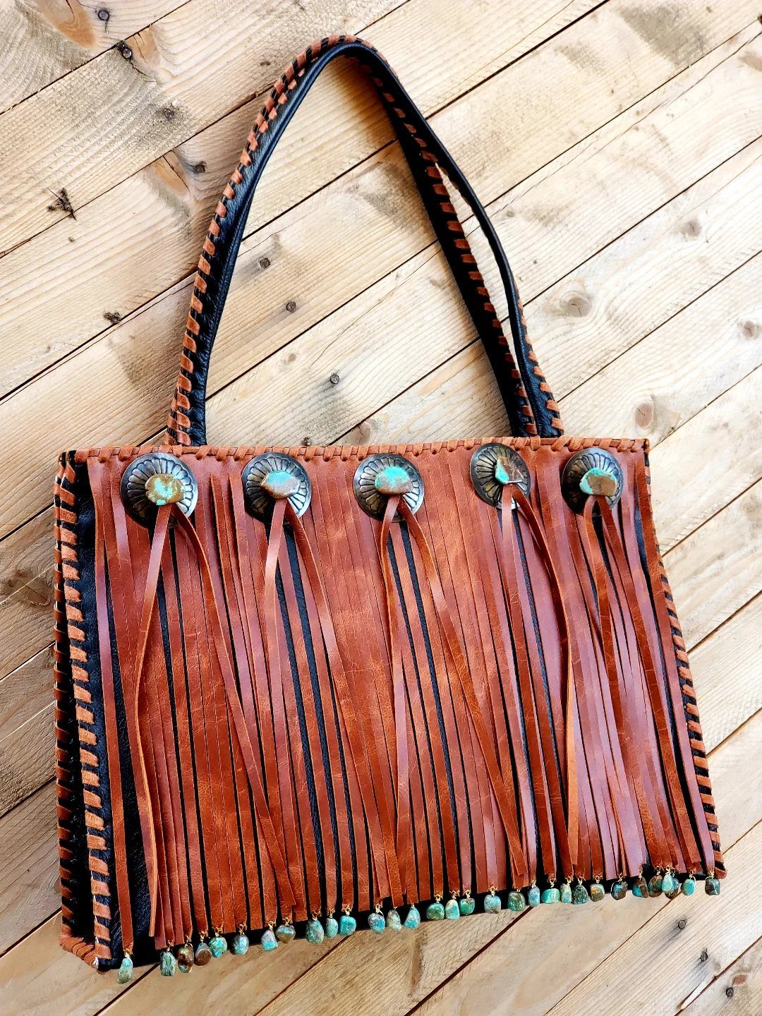 Brown leather fringed handbag with metal conchos on a wooden plank background