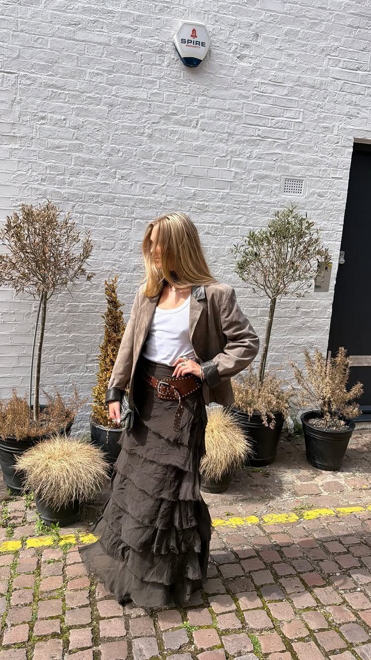 Woman wearing a brown tiered maxi skirt and blazer against a white brick wall.