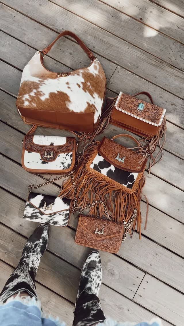Cow-print and brown leather handbags, including one with long fringe, on wooden decking.