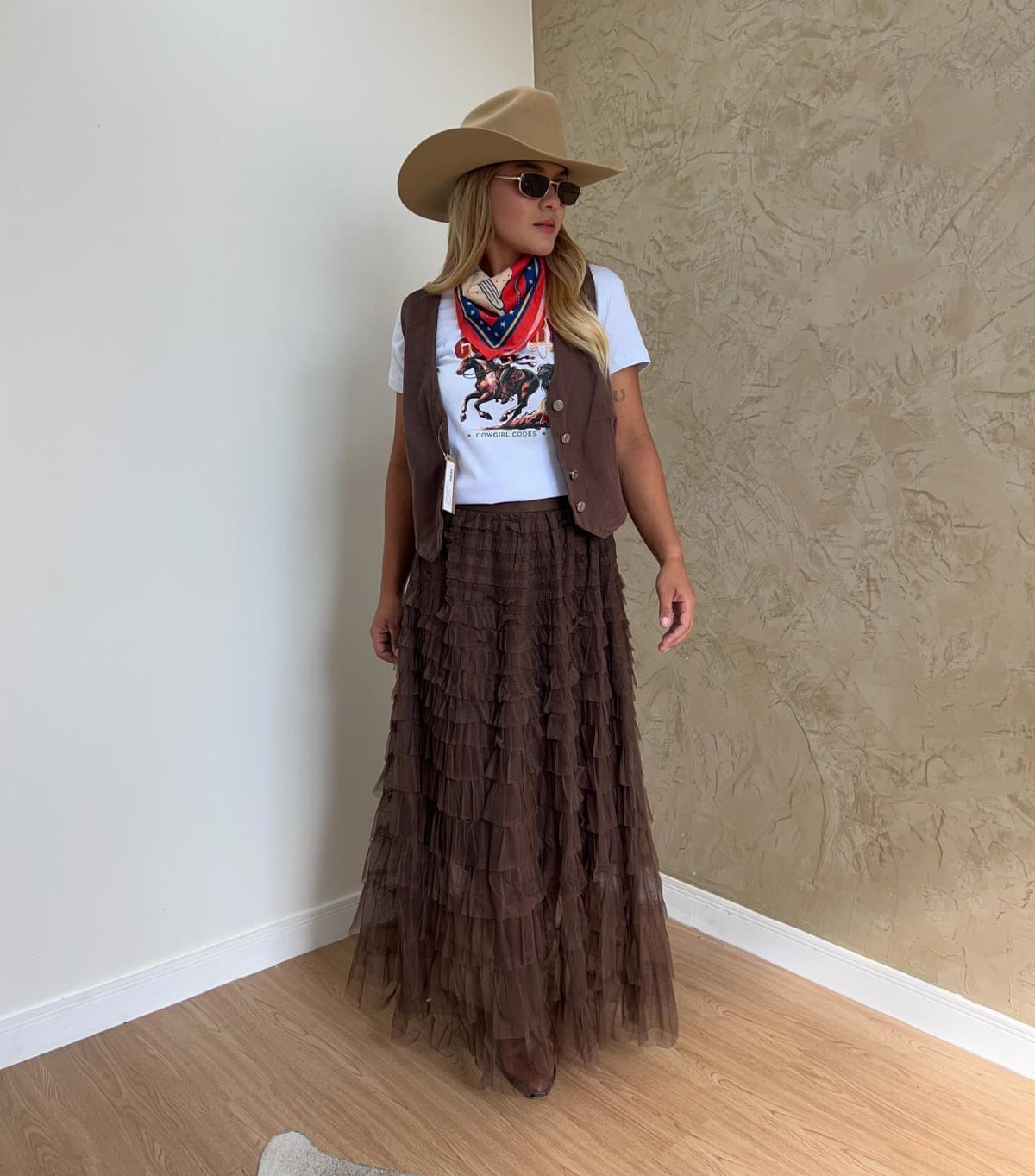 Woman in cowboy hat, graphic tee, brown vest, and tiered ruffled skirt.