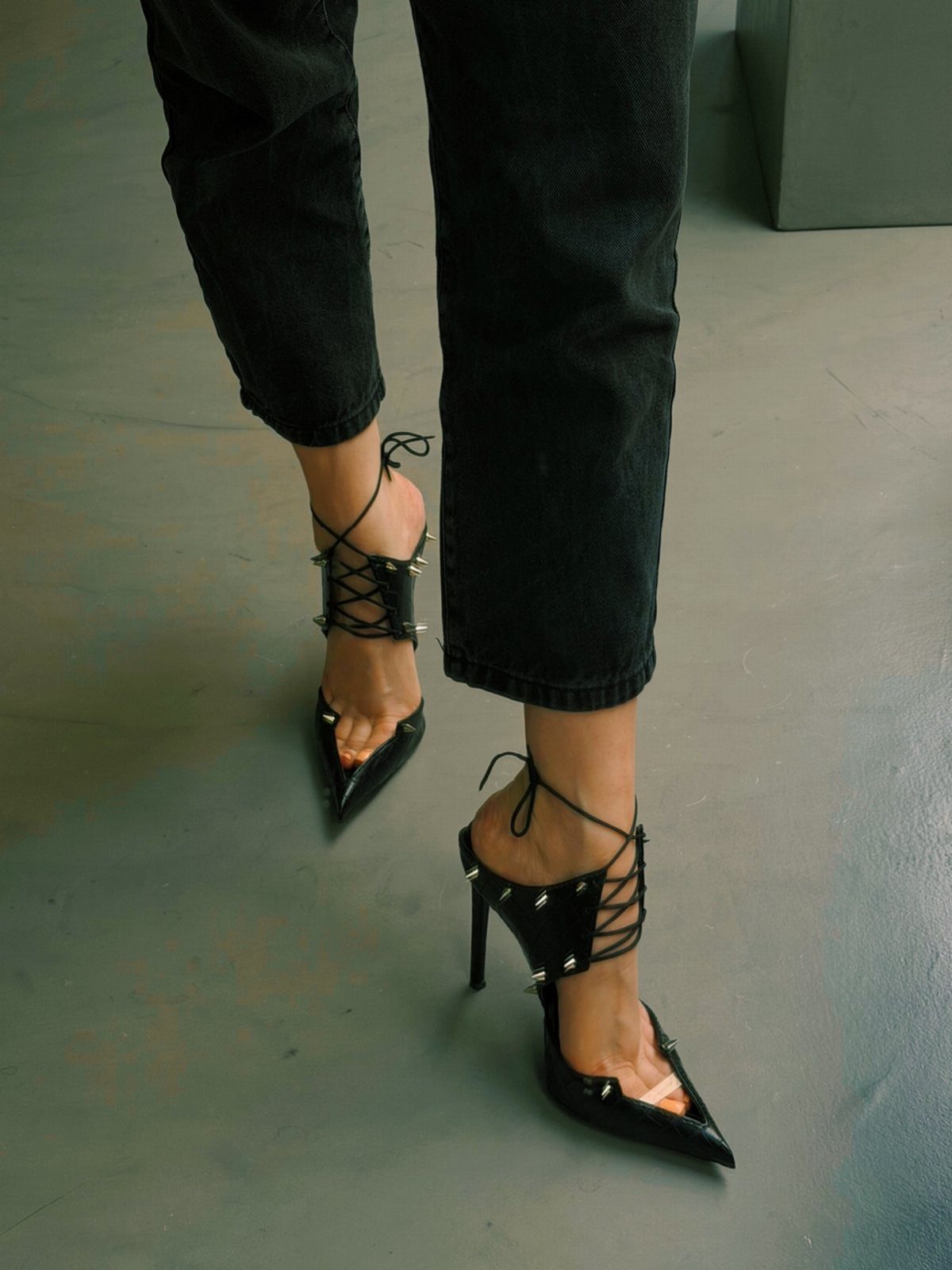 Black pointed-toe high heels with lace-up ankle straps and silver studs on feet.