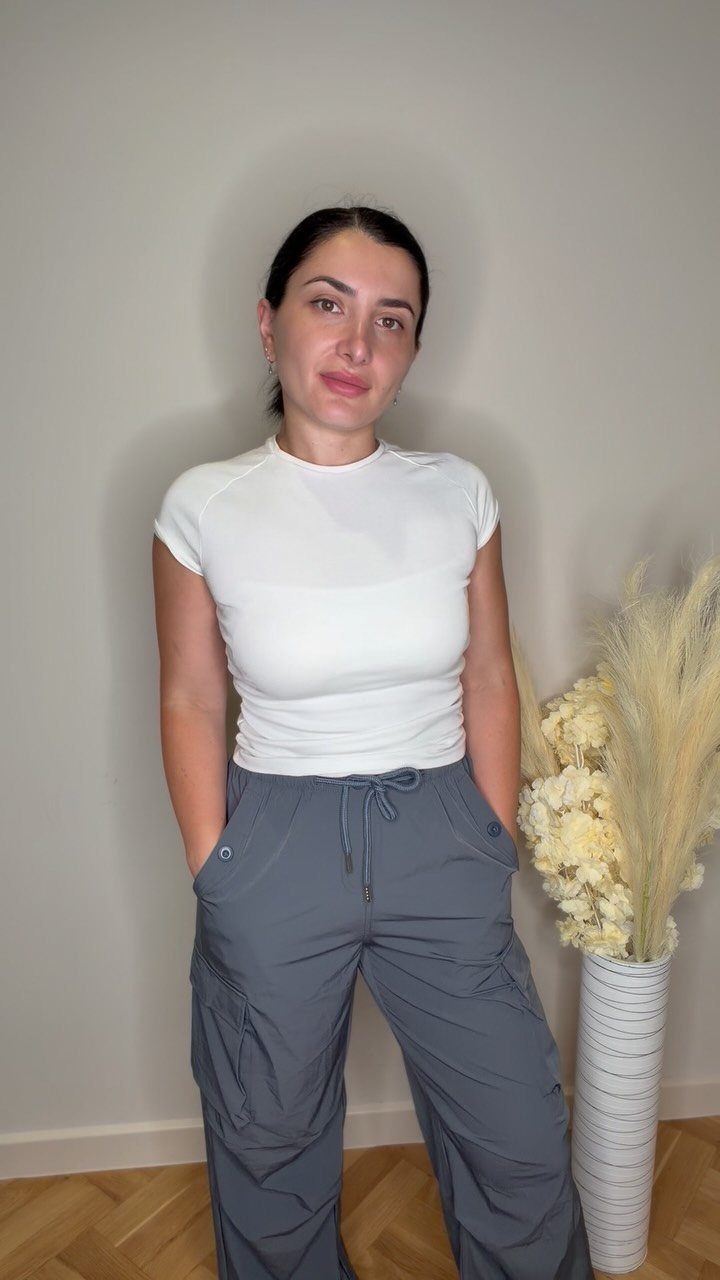 Woman wearing a white fitted t-shirt and baggy grey parachute pants indoors.