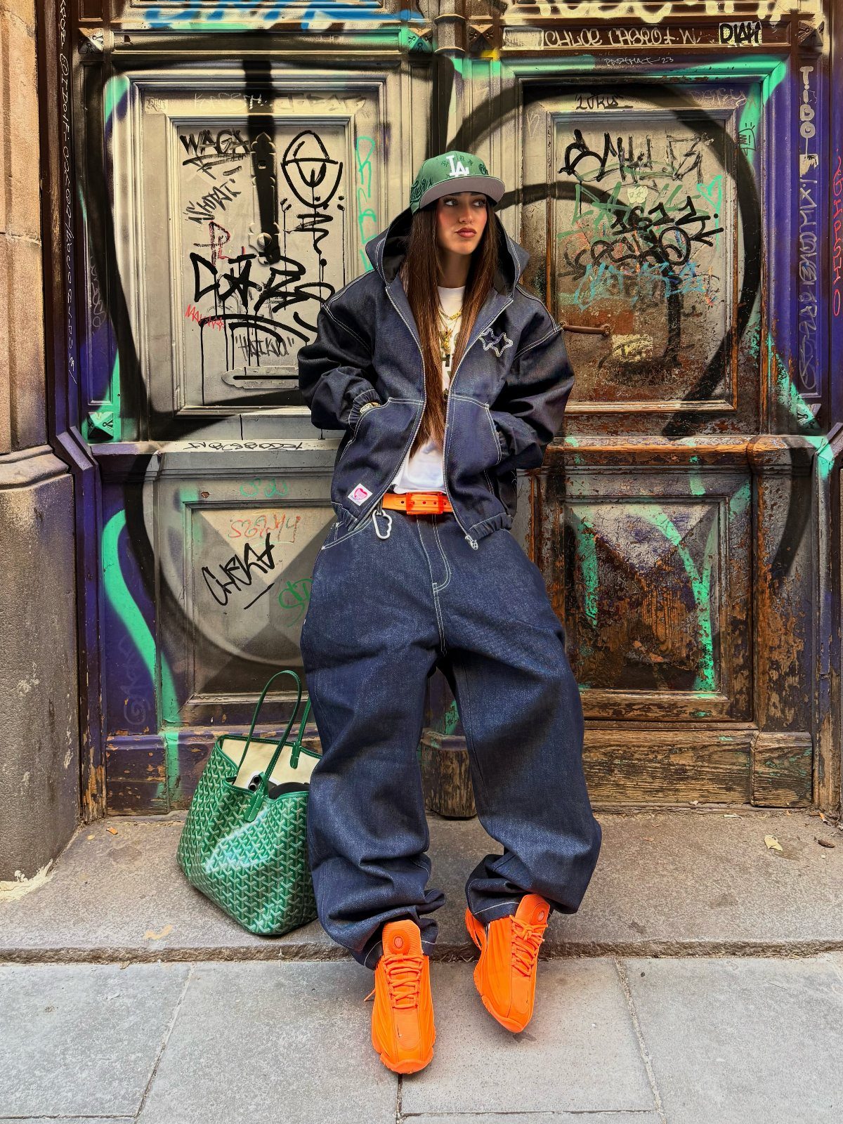 Person in baggy denim jeans, oversized hoodie, and orange sneakers against graffiti wall.