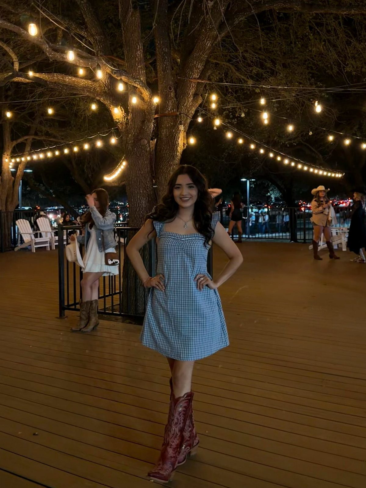 Woman in blue gingham dress and red cowboy boots posing under string lights