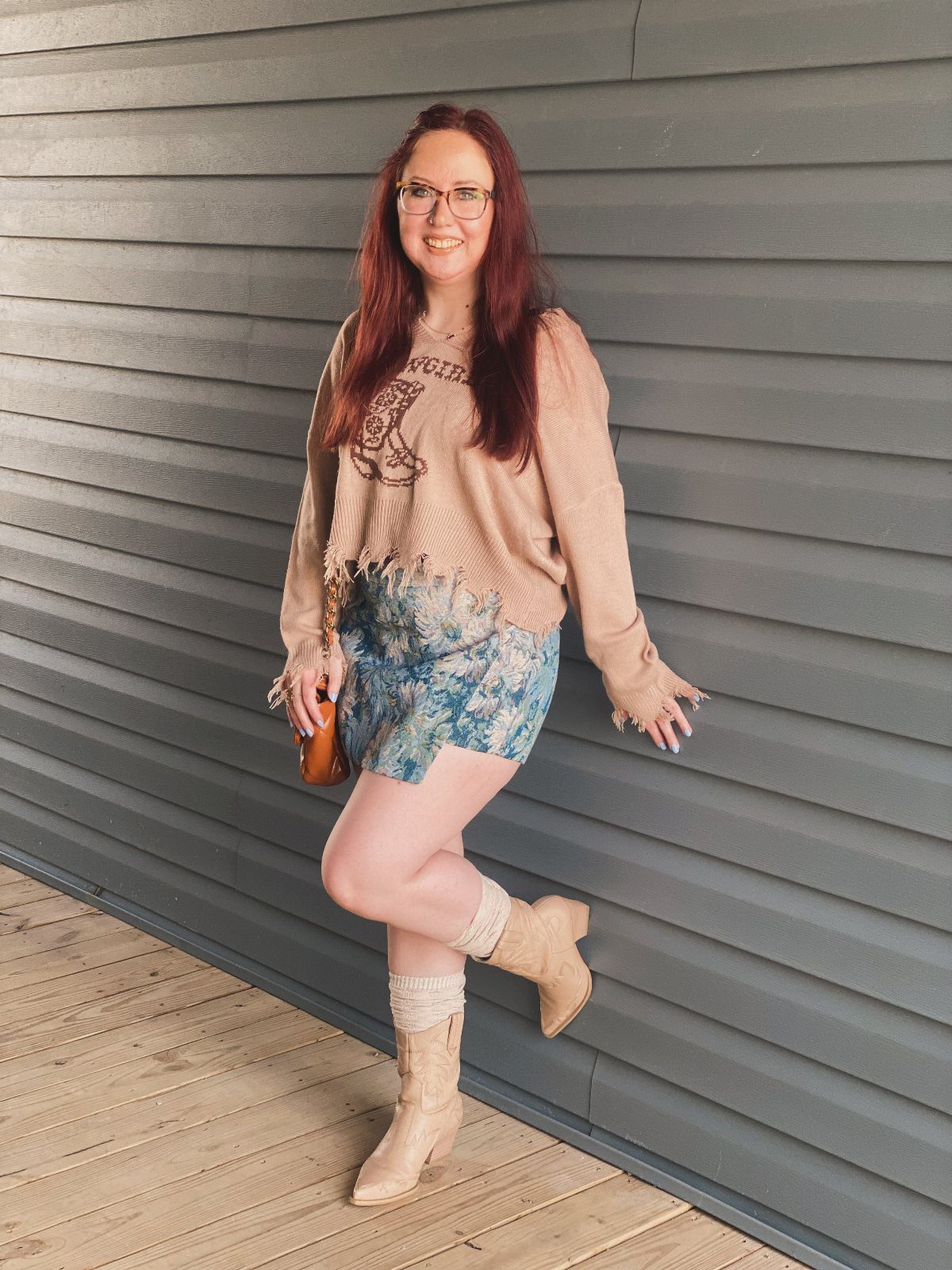 Woman wearing a beige sweater, blue patterned skirt, and beige cowboy boots.